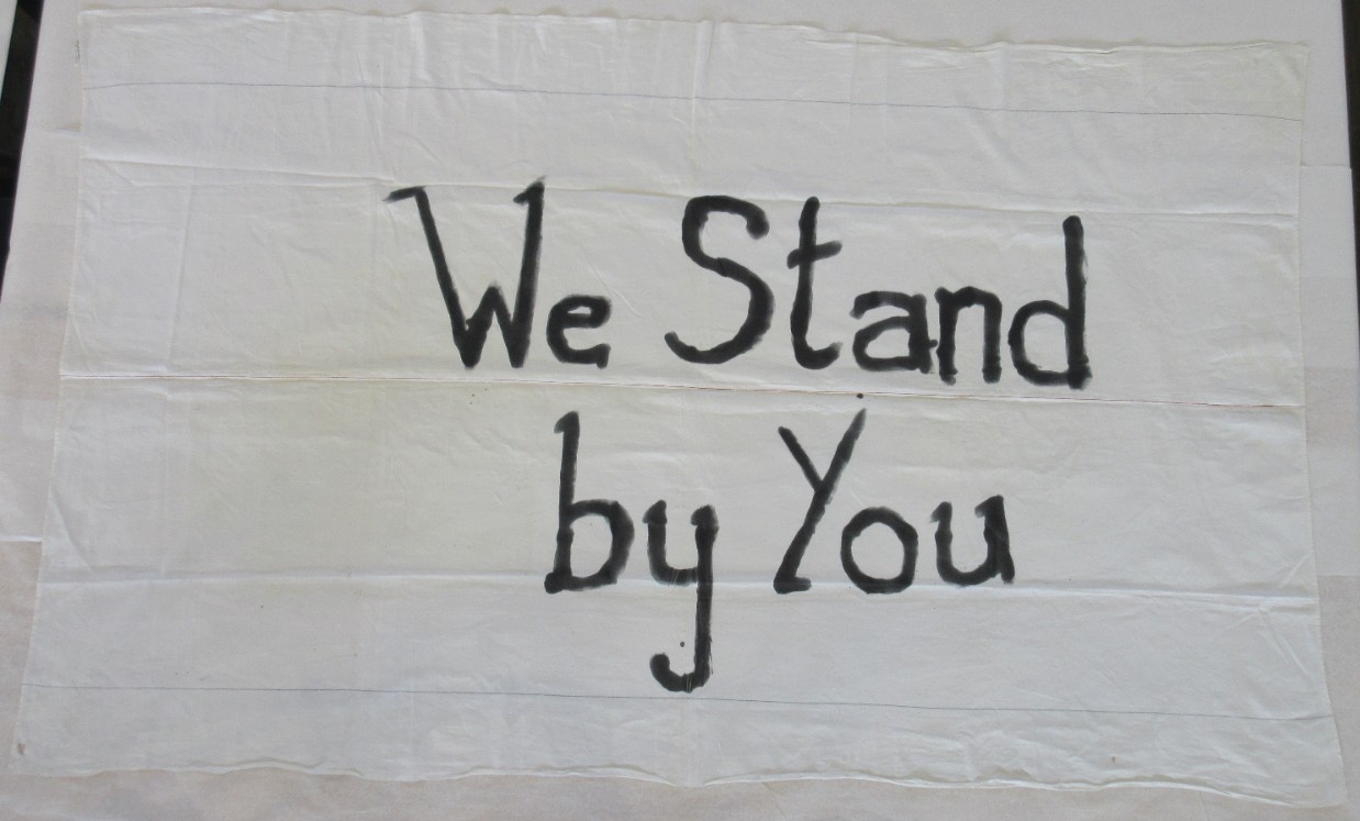 <p>&quot;We Stand by You&quot; written in black ink on white bedsheet.&nbsp;</p>