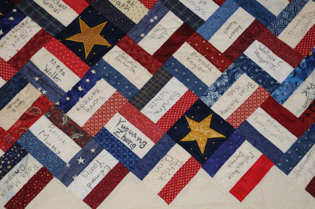 Close-up of 9/11 Pentagon memorial quilt. Quilt consts of red, white, and blue quilting blocks. Names of those who died handwritten on white blocks. Interspersed with blue blocks with gold stars.