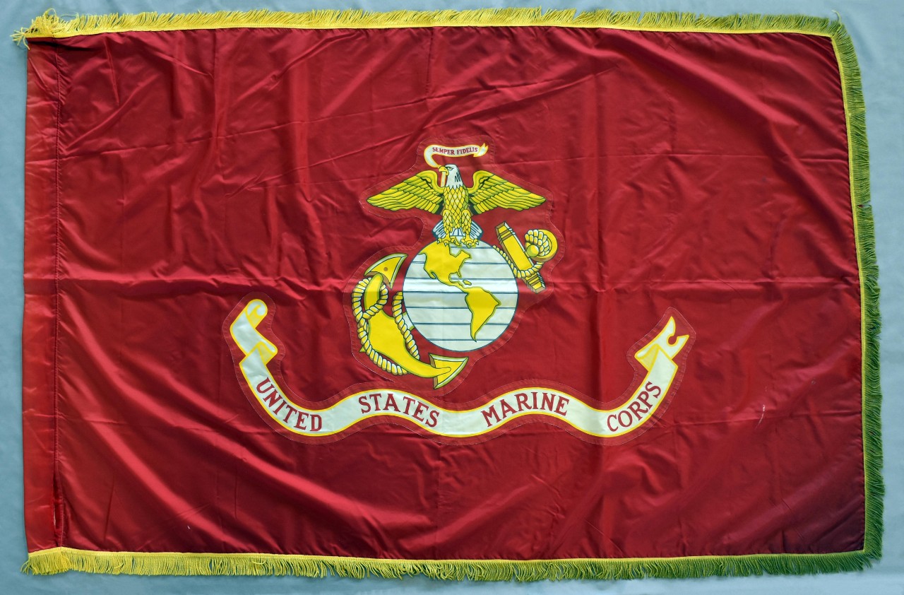 One US Marine Corps flag. The flag is rectangular-shaped red nylon edged with gold fringe. At the center of the flag is the seal of the US Marine Corps with the eagle, globe, and anchor above a banner with the words “United States Marine Corps.” ...