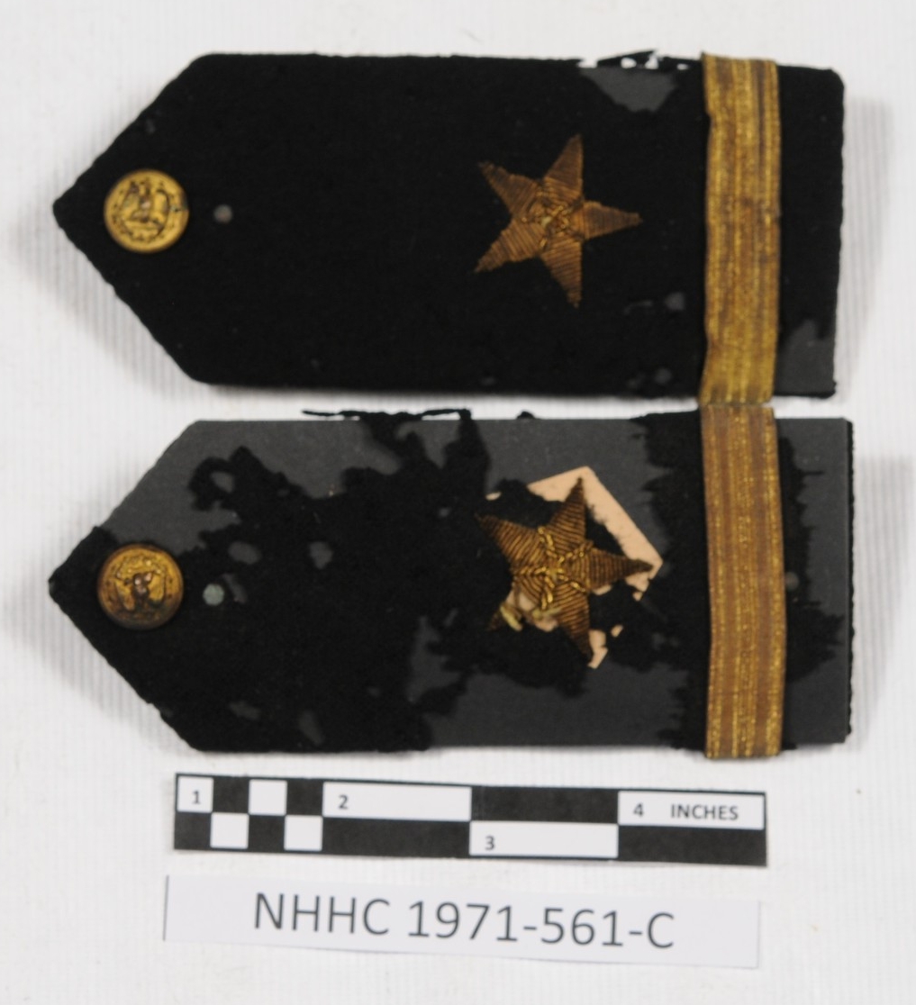 One pair of ensign shoulder boards. Shoulder boards are rectangular in shape, coming to a point at one end. They are covered in black fabric. There is a small gold US Navy button at the point. There is one gold embroidered star and one gold ribbo...