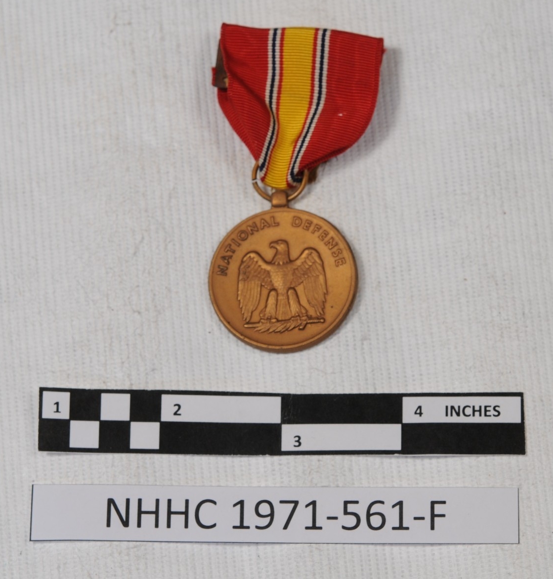<p>One National Defense Service medal. The ribbon has the following vertical stripe color pattern: wide red, thin white, thin dark blue, thin white, thin red, thicker yellow, thin red, thin white, thin dark blue, thin white, wide red. There is a metal backer with a pin to attach the ribbon to a board. The bronze medal is circular. The obverse design has NATIONAL DEFENSE across the top above an eagle with outstretched wings, standing on a sword and a palm branch. </p>
