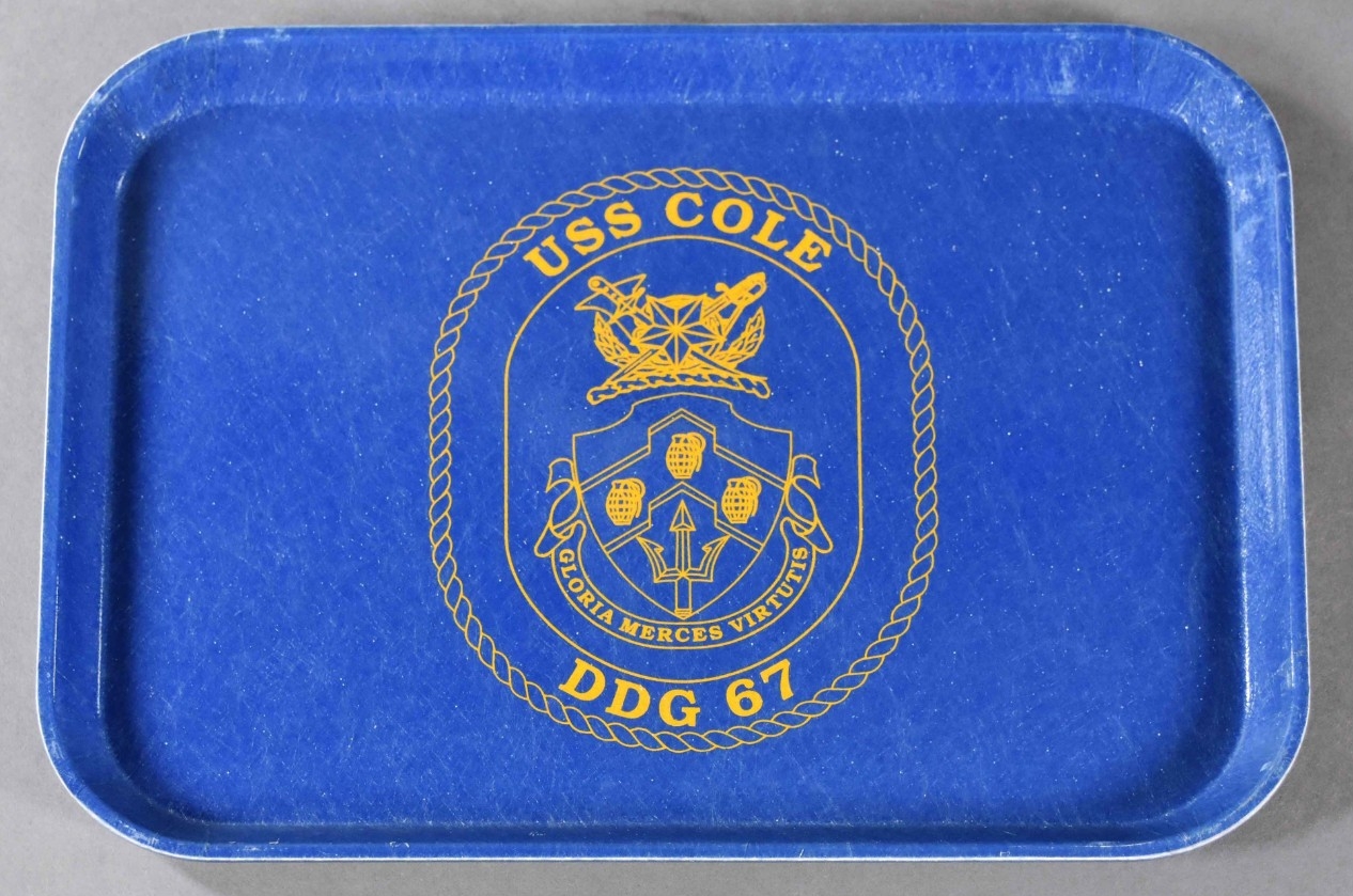 One mess deck tray from USS Cole. The tray is rectangular-shaped blue plastic with raised edges and curved corners. In the center of the obverse side is a gold colored USS Cole (DDG-67) insignia.  