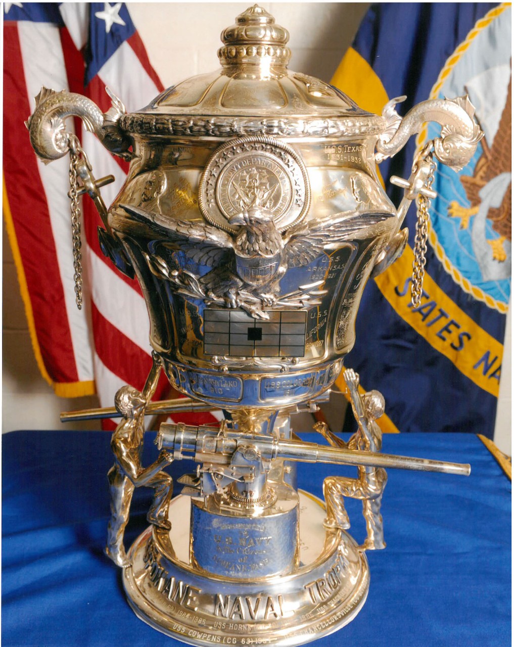 Silver urn style trophy with navy guns and men holding it a target and handles with a lid to cover the cup