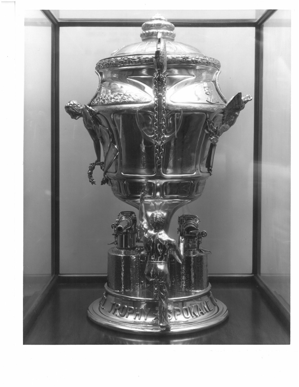 Silver urn style trophy with navy guns and men holding it a target and handles with a lid to cover the cup image of side of cup