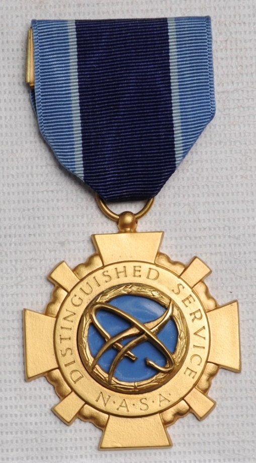 Cross shaped gold colored medal superimposed over a wreath. The center is blue enamel with a 3D NASA insignia on top. In a ring around the center, engraved words read, "DISTINGUISHED SERVICE/NASA". Ribbon is navy blue, and sea blue.