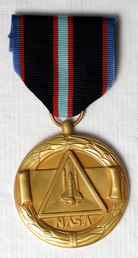 The NASA Space Flight Medal planchet is a wreath with a scroll in the center. in the foreground is a triangle with a space shuttle in the center. below the triangle is the "worm" style NASA logo. The medal has a finger attached to a suspension ri...