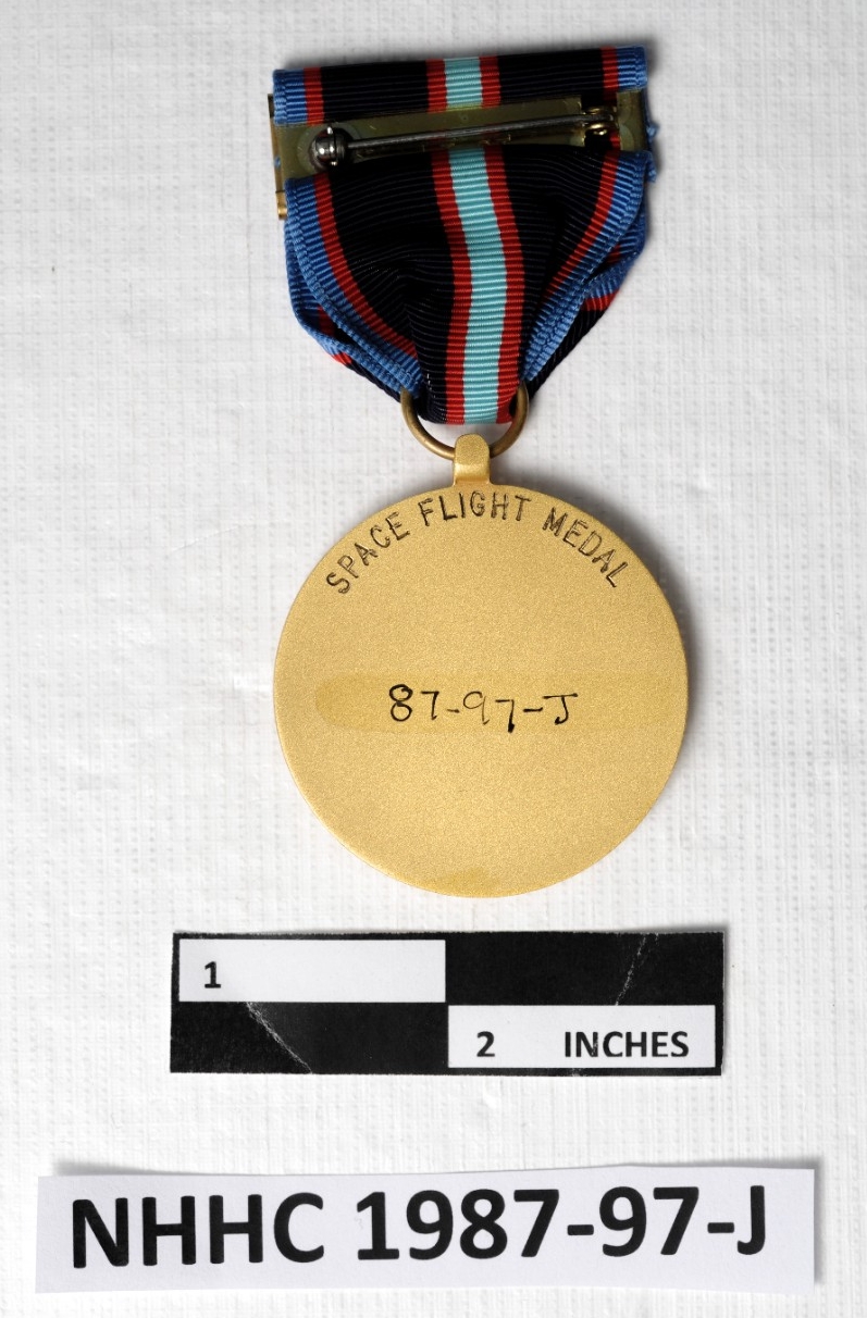 The reverse is stamped "Space Flight Medal" The medal has a finger attached to a suspension ring. The ring taperes the ribbon. The ribbon colors are blue, red, black, red, aquamarine, red, black, red, blue. The pin and clasp is slip on style.  