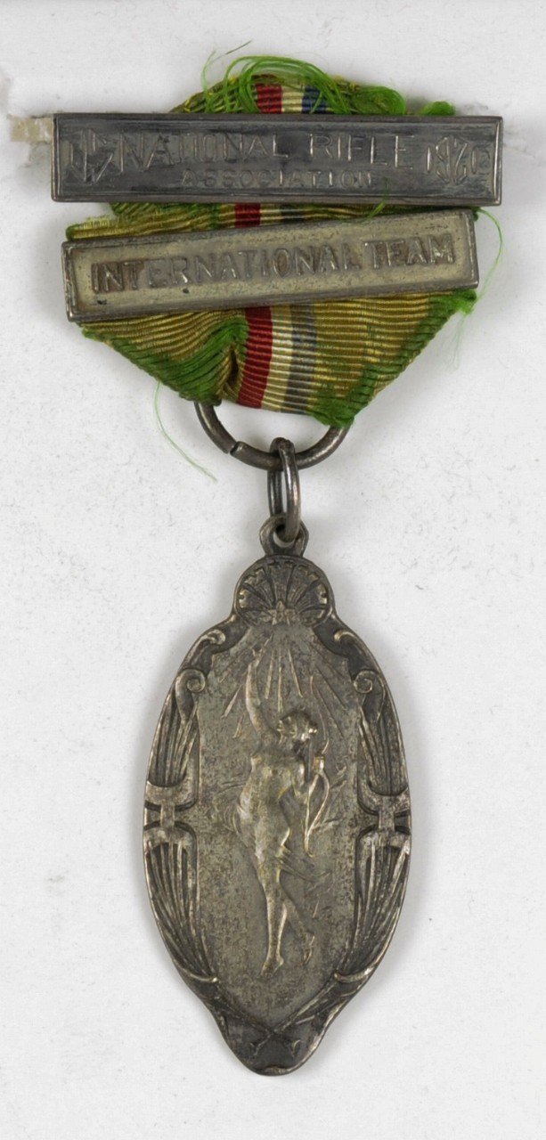 MEDAL-NRA INTERNATIONAL TEAM, UNION INTERNATIONALE DE TIR, MATCHES LYON 1921. THE MEDAL RIBBON IS GREEN WITH A CENTRAL STRIPE OF RED, WHITE AND BLUE. THE MEDAL HAS A BAR/PIN AND ONE ADDITIONAL BAR ON THE RIBBON. THE PLANCHET IS OVAL IN SHAPE. THE...