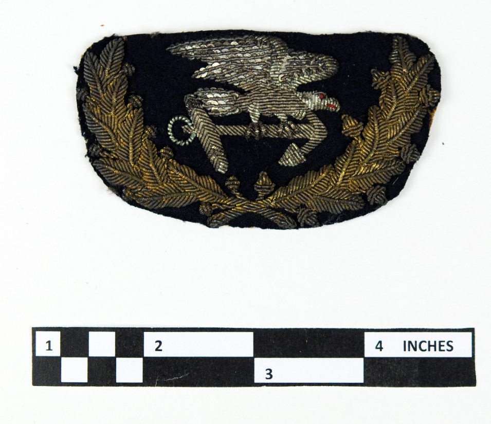 one trapezoidal shaped black wool cap device with gold oak and laurel branch and a silver sequined eagle with red eye and mouth holding an anchor and done in a goldwork style.