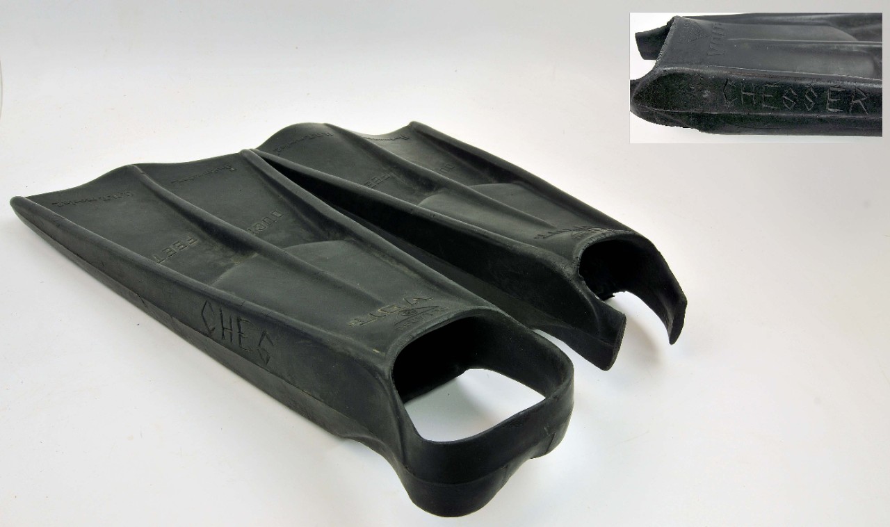 One pair of black hard rubber swim fins. The swim fins are shaped like duck feet. There is a cutout at the back of each fin for a foot to slide in and a black strap to hook around the wearer’s heel. The heel strap is missing a section from one of...