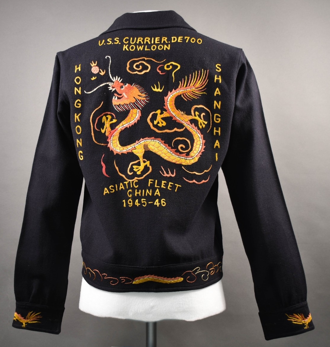 <p>The back of the blue wool cruise jacket decorated with colorful embroidery. At the center of the back is a large colorful embroidered Asian-style dragon. Embroidered in yellow around the edges of the image is “U.S.S. Currier. DE700 / Hong Kong / Kowloon / Shanghai / Asiatic Fleet China 1945-46.” The waist trim is also embroidered with a section of a dragon body and various geometric designs.</p>
