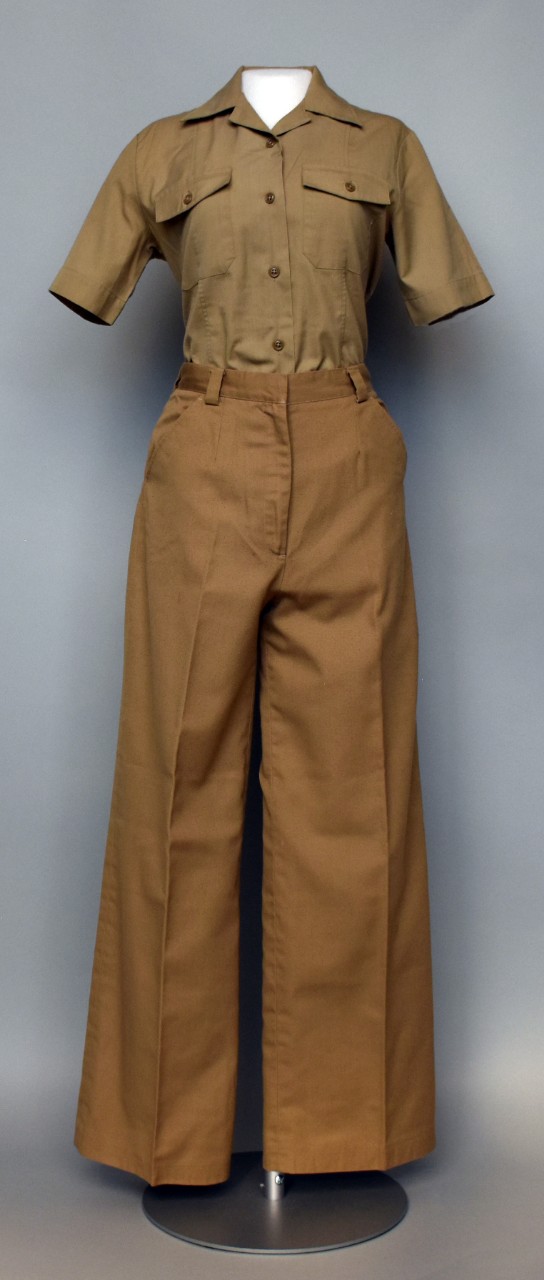 One female Midshipman Service Khaki uniform. Uniform consists of a pair of khaki trousers and a khaki shirt. The trousers have a zipper and hook fly, four pockets, and belt loops around the waist. The shirt has five brown plastic buttons down the...