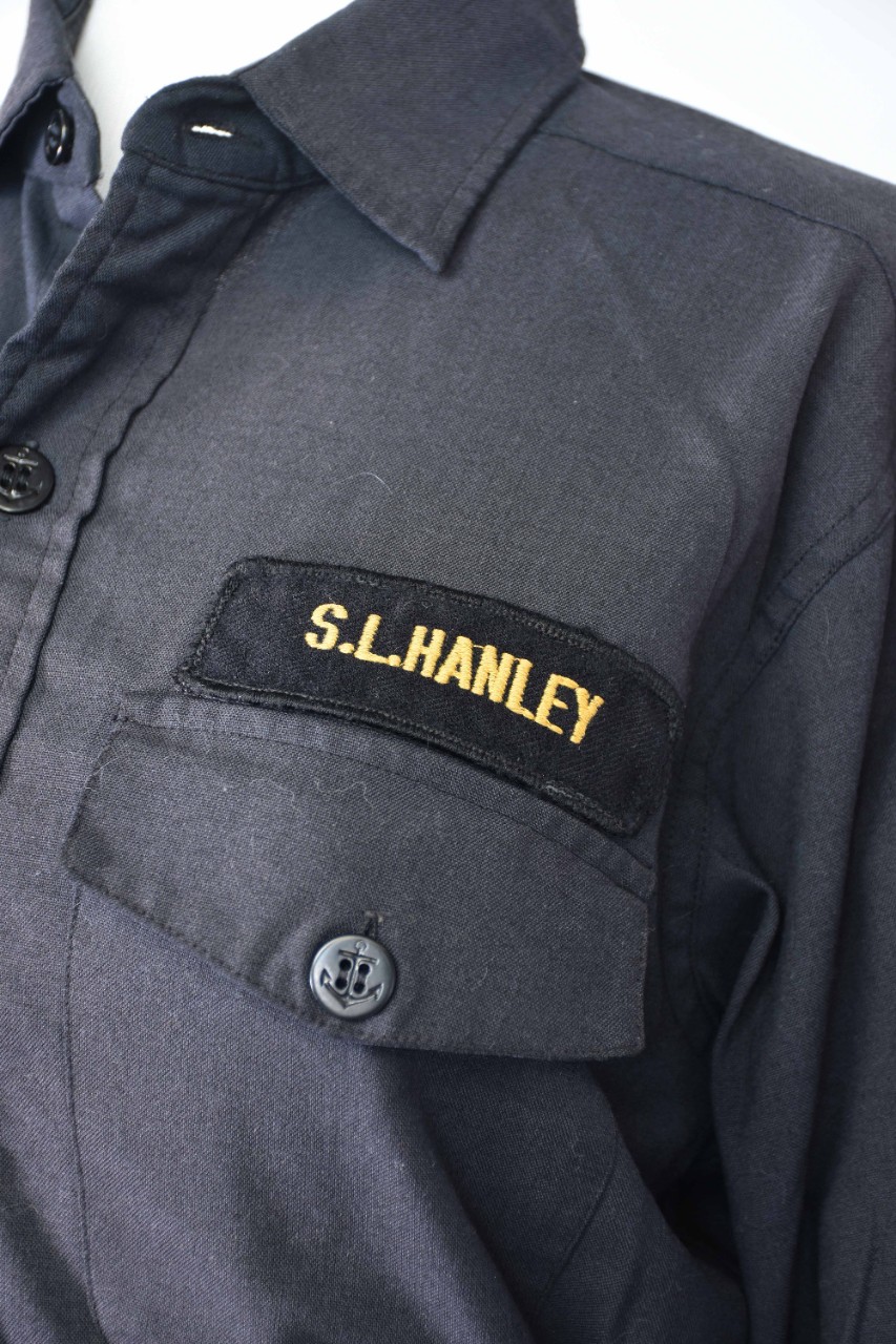 Close-up of name badge on proper left chest above chest pocket of a dark blue uniform shirt. Badge is black fabric embroidered with "S.L.Hanley." 