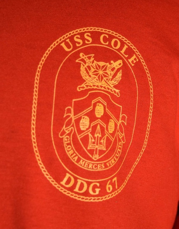 <p>Close-up image of the ship insignia on the front left breast of the food service attendant t-shirt. The insignia is gold printed on red fabric. The insignia is oval-shaped with a rope border. Within the border are the words “USS Cole / DDG 67.” At the top of the insignia is a five-pointed star superimposed on two crossed swords, a US Marine Corps officer sword on the left and a US Navy officer sword on the right. The swords are superimposed on a hunting horn with two laurel branches with a short rope beneath it. Below is a shield with three hand grenades above a trident. Below the shield is a banner with the words “Gloria Merces Virtutis.”</p>