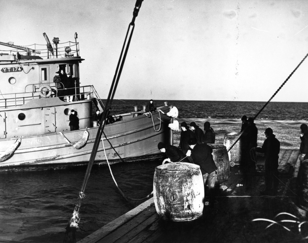 USS Allaquippa (YT-174) mooring to a barge, in Long Island Sound, during net-laying operations in January 1943. Note sailors' cold weather gear, buoys in foreground (on barge), ship's number board on deckhouse, and covered .30 caliber Lewis machine gun atop tug's pilot house.