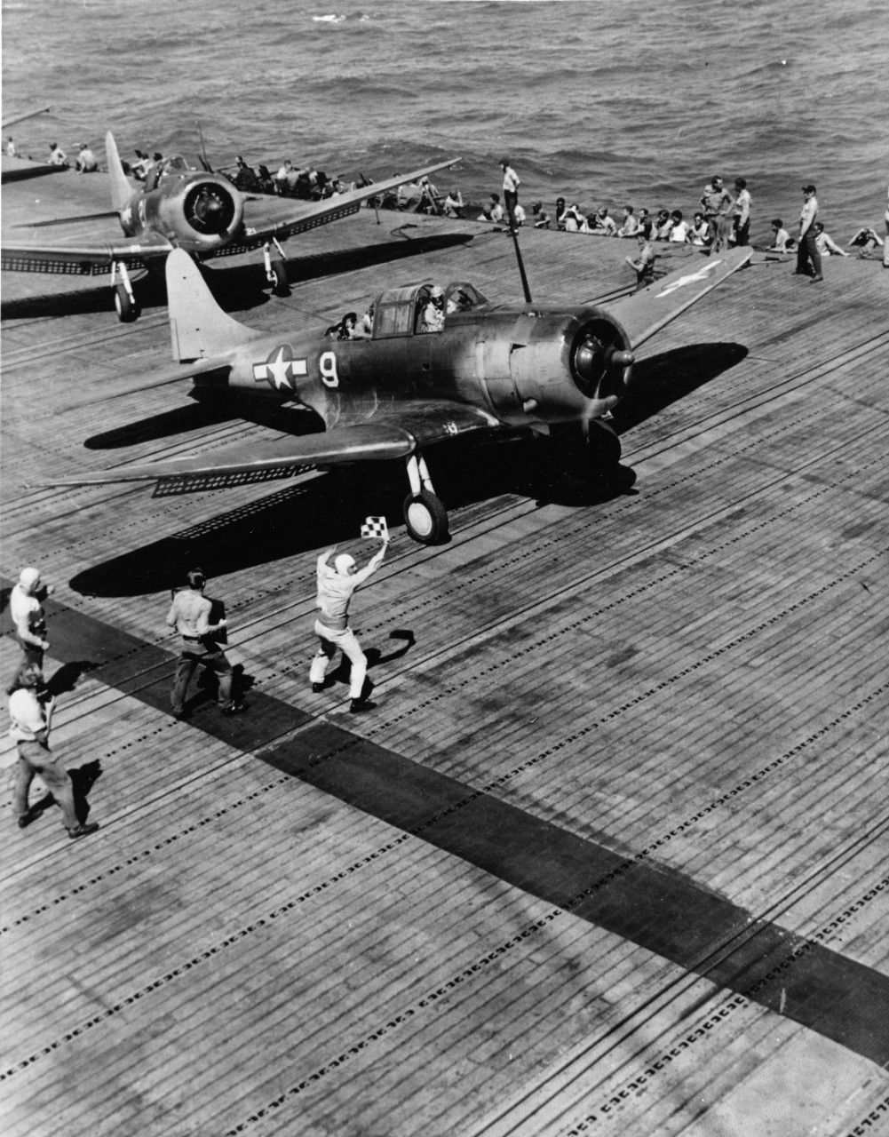 Douglas SBD-5 on the flight deck of USS Yorktown (CV-10) awaits the take-off signal during a raid on a Japanese base, 1 September - 6 October 1943. Note flight deck officer holding flag, and man holding sign with last minute instructions for the pilot.