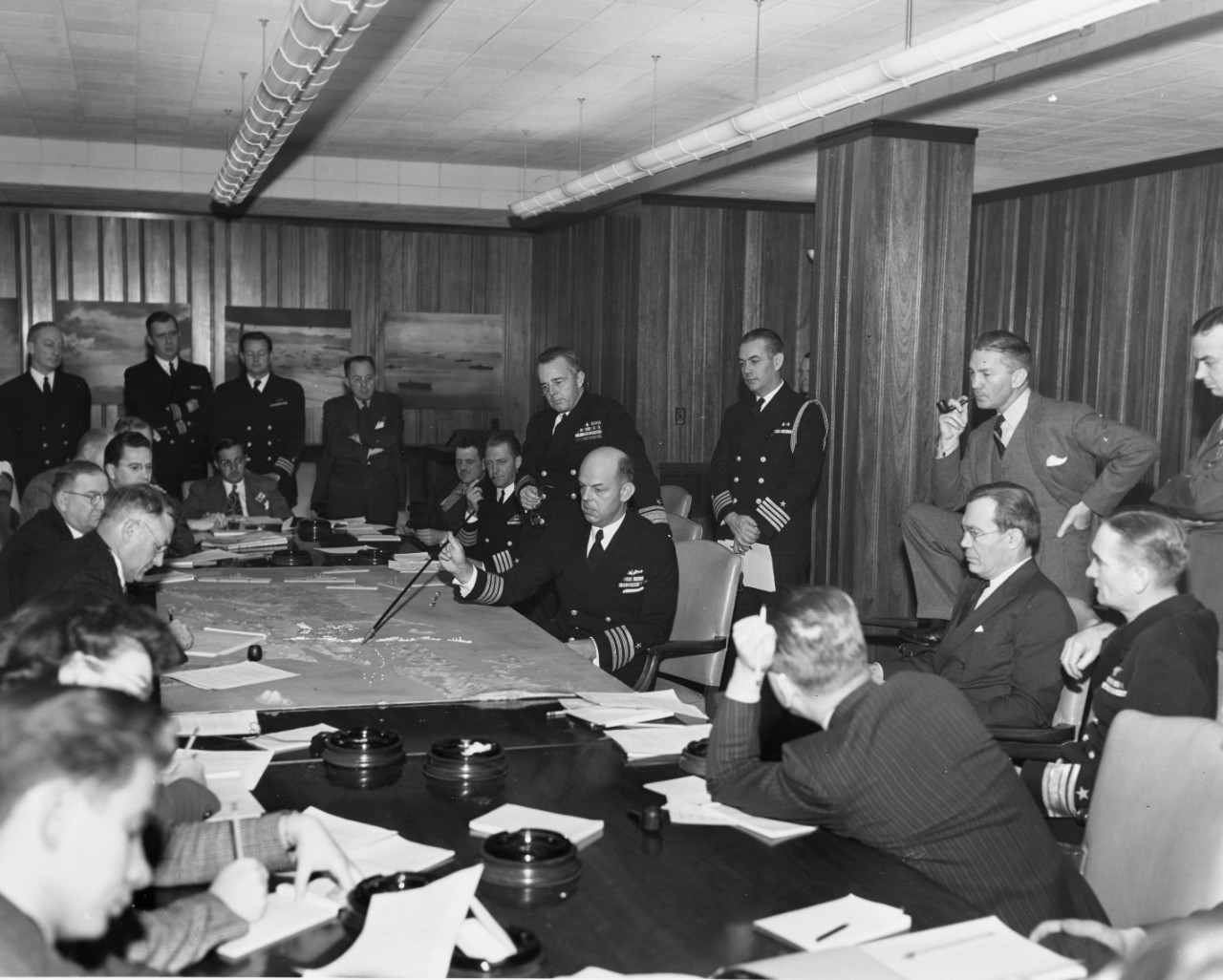 Captain Kenmore M. McManes, USN, describing the Surigao Strait battle of 24 October 1944, to Secretary of the Navy James Forrestal (standing, with pipe) and others in Washington, December 1944.
