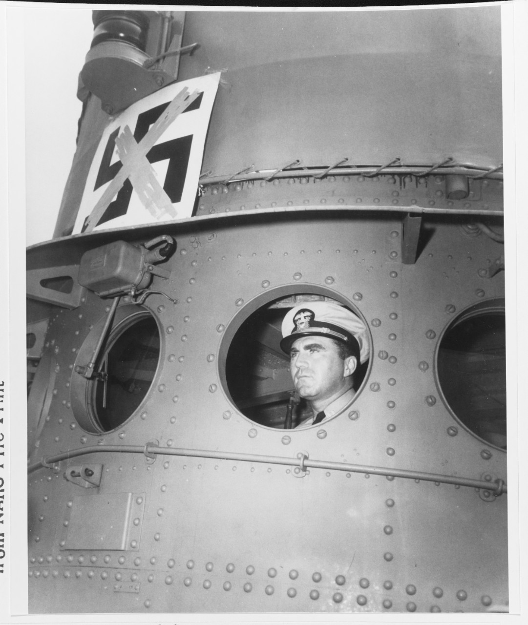 Ship's commander officer, LT Walter T. Flynn, USNR, looks out from a pilot house porthole, 9 August 1943. His ship sank German submarine U-521 off the US East Coast on 2 June 1932. Symbol on bridge face commemorates the event. Note riveted plating, and porthole wiper. 