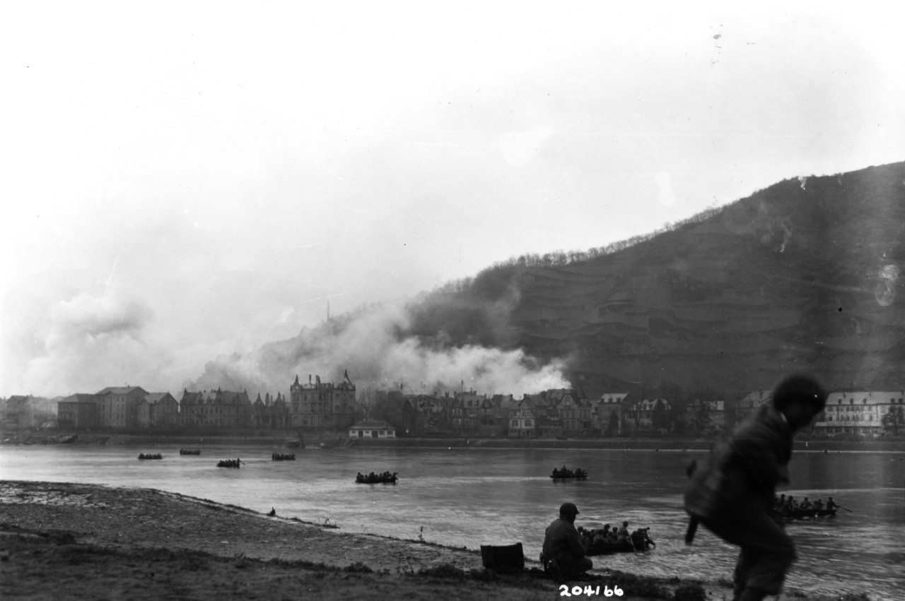 Assault boats carry US Army troops across the Rhine River at St. Goar, Germany, 26 March 1945. Troops on the near shore are covering the crossing against snipers. Note fires burning in town on the opposite bank.
