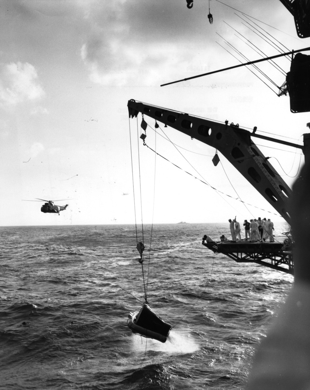 The Gemini 3 capsule is hoisted from the Atlantic Ocean by a large crane on board USS Intrepid (CV-11). Note the helicopter in the background, likely from squadron HS-3.