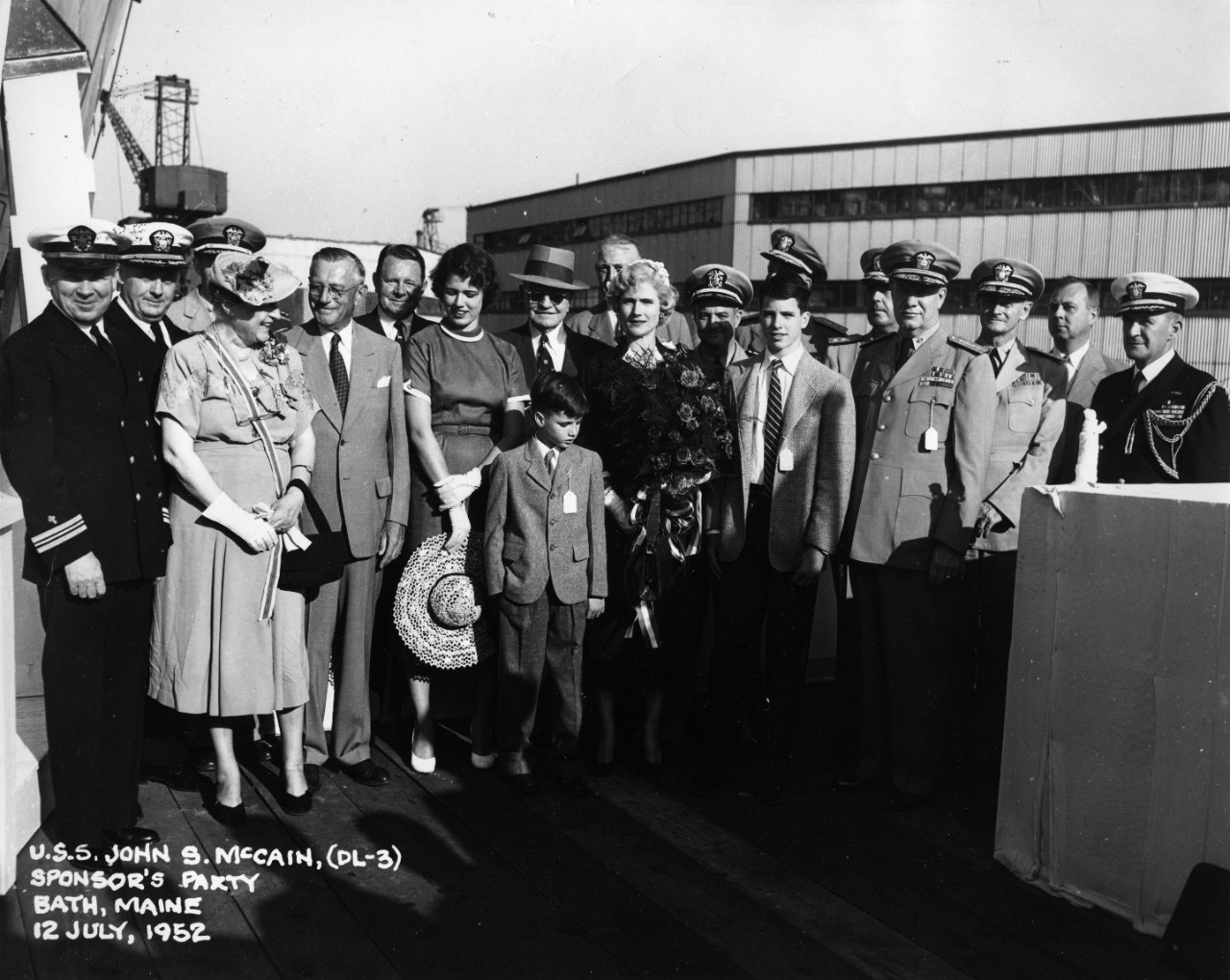 USS John McCain (DL-3) Sponsor's Party, at Bath, Maine. Ship's sponsor Roberta McCain is at center, with her husband Captain John McCain, Jr. behind her in shadow. Her son John McCain III is to their right. To the left, wearing a hat and sunglasses, is retired Fleet Admiral William Halsey.