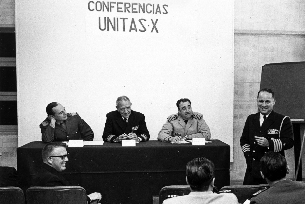 Seated at the table before a UNITAS X pre-sail conference are (left to right) Captain Victor H. Pereyra Murray, Argentine Navy; Rear Admiral James A. Dare, US Navy; and Captain German Da Costa, Uruguayan Navy. Captain Alan J. Kaye, US Navy (right) addresses the conference in Puerto Belgrano, Argentina, October 10. 