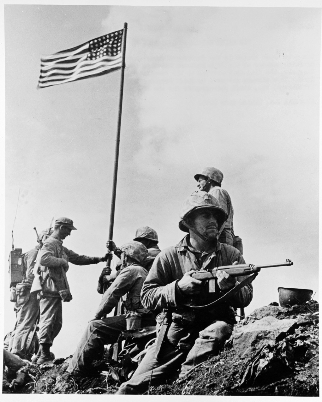 Photo #: NH 104150 Iwo Jima Operation, 1945. The First Flag Raising on Mount Suribachi, Iwo Jima, 23 February 1945 Marines of the 28th Regiment, Fifth Marine Division, hoist the U.S. flag on a piece of pipe, at about 1020 Hrs. on 23 February 1945, after they had captured the summit of Mount Suribachi. This was some seventeen minutes before the famous flag-raising immortalized by Associated Press photographer Joe Rosenthal. Holding the flagpole are Sergeant Henry O. Hansen, Private Philip L. Ward, Pharmacist’s Mate 2<sup>nd</sup> Class John Bradley. In the foreground Private First Class J.R. Michaels stands guard with an M-1 Carbine. Corporal C.W. Lindberg is behind him. (details from Morison: Vol. XIV, frontispiece and page 61). Taken by Staff Sergeant Louis R. Lowery, USMC, staff photographer for Leatherneck magazine. The original photograph came from the illustrations package for Rear Admiral Samuel Eliot Morison's History of United States Naval Operations in World War II, volume XIV: Victory in the Pacific (frontispiece). Official U.S. Marine Corps Photograph, from the collections of the Naval History and Heritage Command.