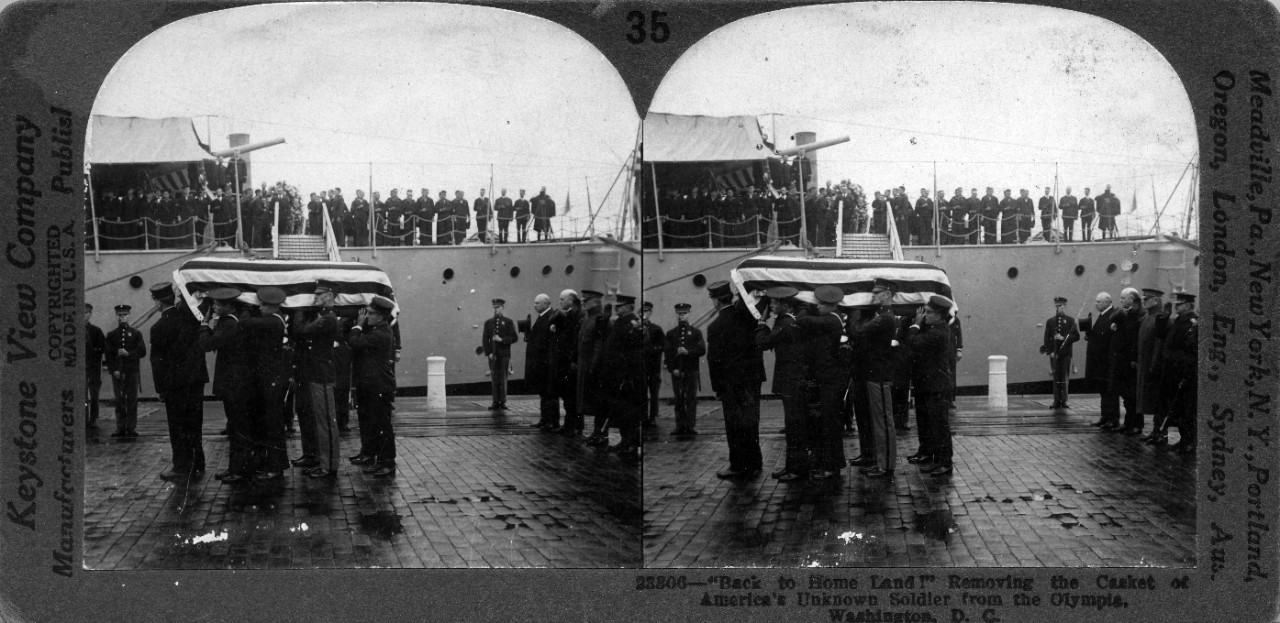 Stereo card showing the casket of the Unknown Soldier being removed from USS Olympia (C-6) at the Washington Navy Yard, returning home from France. At extreme right are (L-R): Secretary of the Navy Edwin Denby, General John J. Pershing, and Admiral Robert E. Coontz. GMC Roy E.B. Larson is the last man at right holding the casket. Originally published by Keystone View Company.