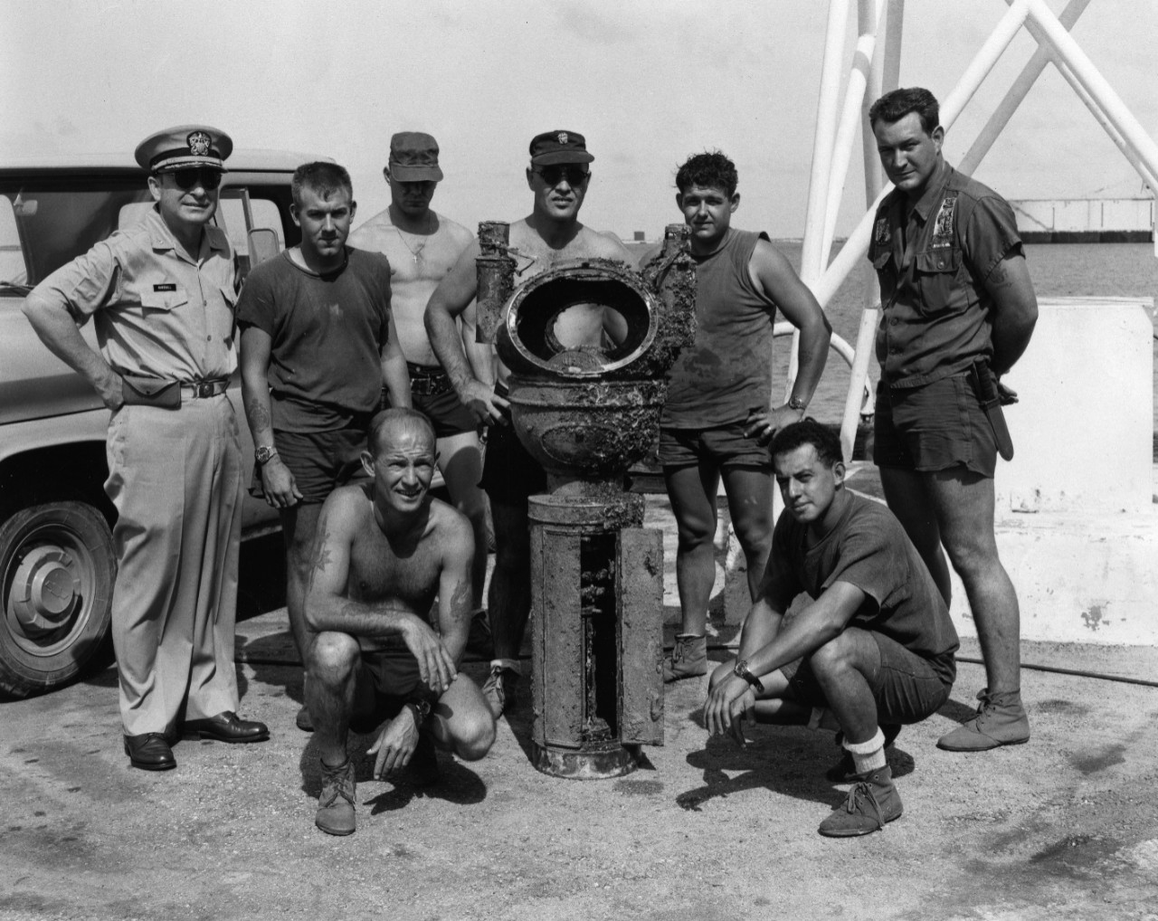 Recovery crew pose with binnacle from SMS Cormoran, salvaged out of Guam Harbor in 1965. Officer at left is Captain W.L. Marshall, Commanding Officer of the Ship Repair Facility. Shirtless individual directly behind binnacle may be the donor, Lieutenant Commander John Naquin.