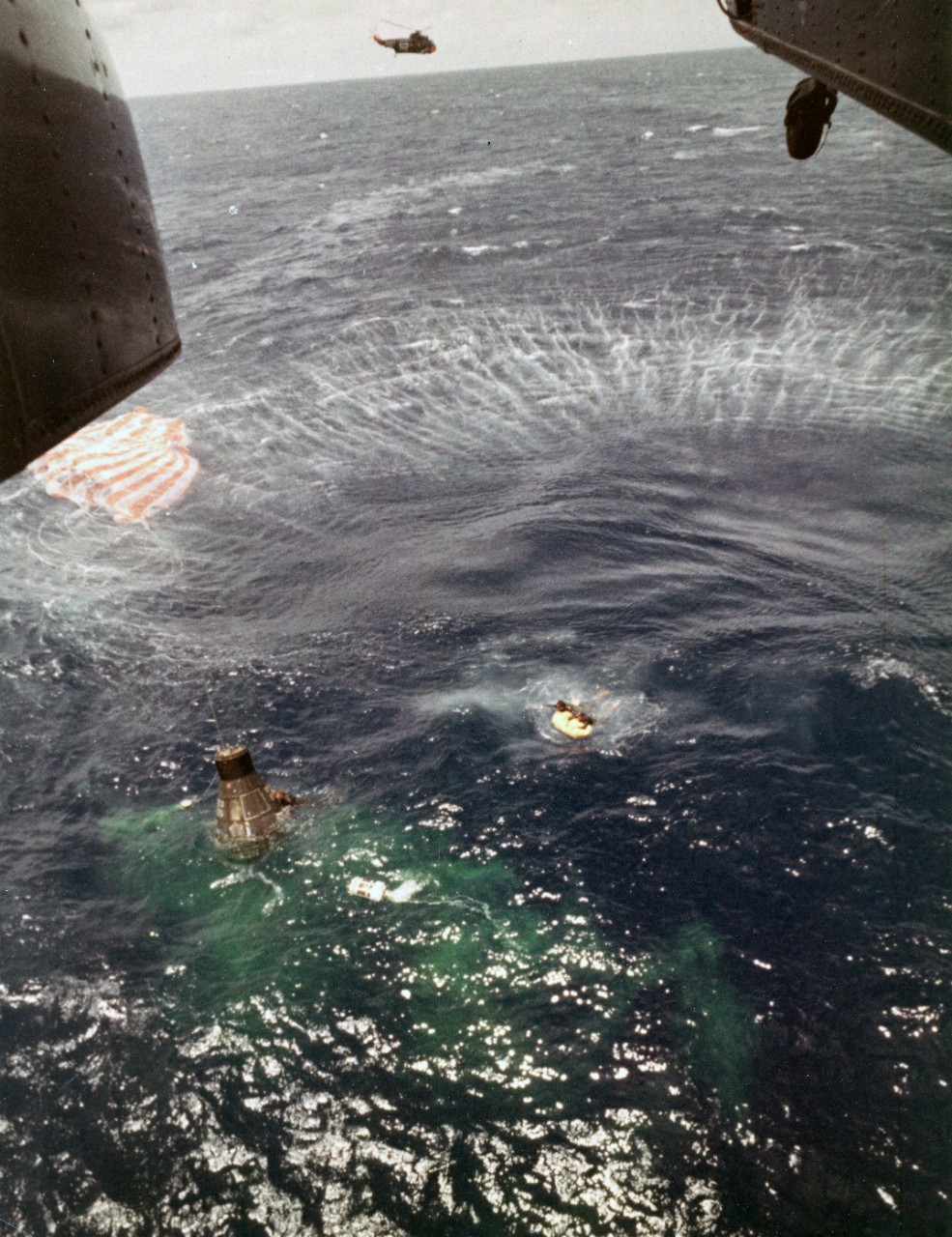 A U.S. Navy frogman, deployed from the hovering helicopter, swims next to the spacecraft and makes contact with Astronaut Cooper inside, as his fellow team members bring up the flotation gear to be attached to the spacecraft. The main chute floats at the top left, and the ejected unused reserve chute floats at the lower right of the spacecraft in the green dye area.