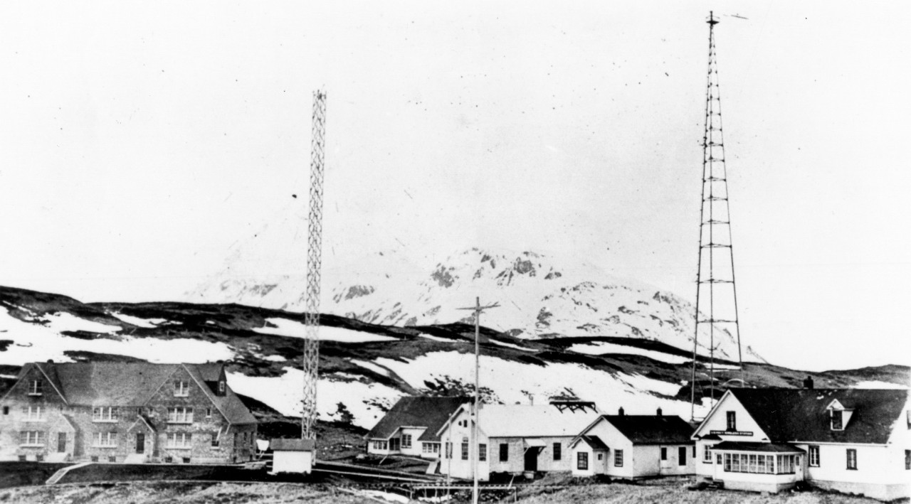 <p>UA 437.04.14 US Naval Radio Station, Dutch Harbor, T.A. Large building of left, apartments for men and families. Receiving and sending room in building directly behind signal mast, with yard in foreground of picture. This station also houses Aerological unit. Large fabricated tower to right of apartment house has been replaced by one of wood. See No. 7 following. Apartment house has large well furnished rooms, well heated. Tramway and bridge leads down to docks in bay. See no. 5 Freight is hauled up to the radio station on cars by stteel cable and power operated windlass. Unalaska only reached by boat from Radio station which now has new motor whale boat (Navy standard).&nbsp;</p>