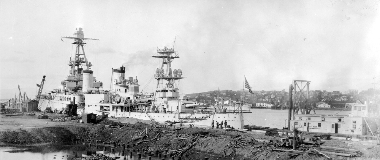 Chicago, tied up alongside ongoing construction of the new 1,000-foot quay wall at Mare Island, 21 February 1934, following repairs necessitated by the collision with Silverpalm. Note that the ship still retains her torpedo tubes at this stage in her life, that an “E” with hash mark is painted on Turret III (as well as on the after stack), and the U.S. Army Corps of Engineers vessel in the stream in the background. (U.S. Navy Bureau of Yards and Docks Photograph, RG 71, Box 71-CA-244-A, National Archives and Records Administration, Still Pictures Branch, College Park, Md.)