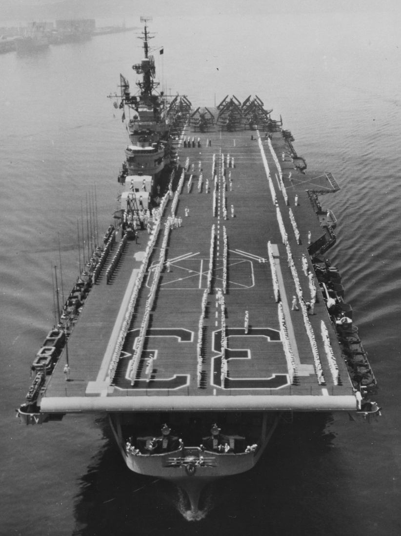 The ship’s company parades for inspection on the flight deck in this bow on view of the ship, late 1940s. Note the gun tubs and the planes parked aft. (Naval History and Heritage Command Photograph 97402)