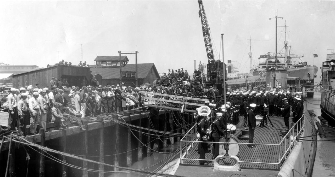 While onlookers of all ages line the nearby pier and other nearby vantage points, an officer – probably Capt. Thomas A. Kearny, the Captain of the Yard – reads the orders placing V-6, her main 6-inch battery not yet fitted, in commission on 1 July 1930 while she lies alongside an overage destroyer being prepared for disposition. Note band and color guard along the port side, and ship’s company lined up athwart ships, while a sailor readies the flag to hoist at the appropriate moment, and the enlisted man at the bass drum glances toward the photographer. (U.S. Navy Bureau of Ships Photograph RG 19-LCM, 19-LC-53-F-4, National Archives and Records Administration, Still Pictures Branch, College Park, Md.)