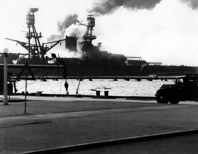 Nevada heading down the channel, afire from several Japanese bomb hits, as seen from Ford Island during the latter part of the attack. Ship whose boom and flagstaff are visible at left is Avocet (AVP-4). Note camouflage Measure 5 false bow wave painted on Nevada. (U.S. Navy Photograph 80-G-32443 National Archives and Records Administration, Still Pictures Division, College Park, Md.)