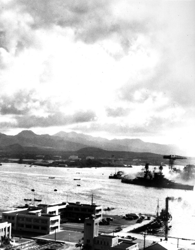 Nevada heading down the channel past the Navy Yard's 1010 Dock, under Japanese air attack during her sortie from Battleship Row. The camouflage Measure 5 false bow wave is faintly visible painted on the battleship's forward hull. Photographed from Ford Island. The smaller ship in the lower right is the tender Avocet (AVP-4). Note fuel tank farm in the left center distance, beyond the Submarine Base. (Naval History and Heritage Command Photograph NH 97397)