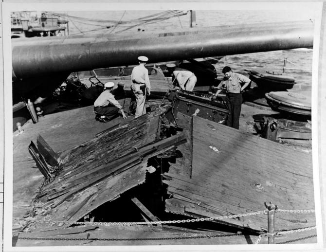 Damage to the forecastle deck of Nevada, caused by the explosion of a Japanese bomb below decks. Gun barrels of the battleship's forward 14/45 triple turret are in the background. Photographed on 12 December 1941 from on board Rail (AM-26), which was tied up alongside Nevada's starboard bow, assisting with salvage efforts. Note officer in center, wearing a .45 caliber pistol. (Naval History and Heritage Command Photograph NH 64484)
