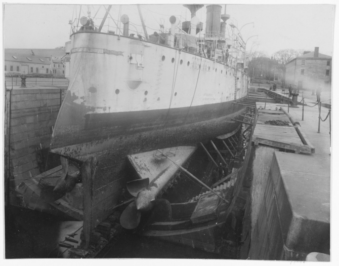 Olympia completes repairs and an overhaul in drydock at Boston Navy Yard, Mass., c. November–December 1899. Note her nameplate, and the badly damaged starboard propeller. (Unattributed U.S. Navy Photograph NH 43334, Photographic Section, Naval History and Heritage Command)