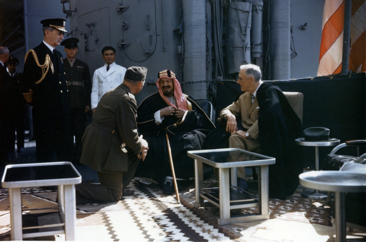 Roosevelt and King Saud confer on board Quincy, 14 February 1945. The monarch speaks to Col. Eddy, who interprets the conversation, and Fleet Adm. Leahy is at left. In addition to the stately furniture, an ornate carpet is draped upon the ship’s ...