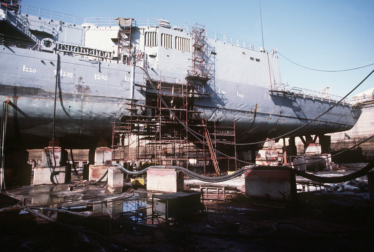 The size of the hole bears mute testimony to the fury of the mine’s detonation, while Samuel B. Roberts rests in dry dock at Dubai in the United Arab Emirates. Workers clamber over scaffolding and temporarily make the ship seaworthy for her voyage to the United States. (PH1 Chuck Mussi, All Hands, August 1988, No. 857, page 14)