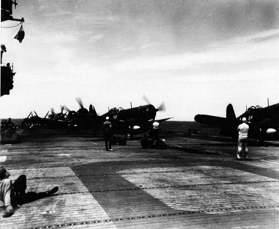 The ship continues her war effort as she launches the Corsairs into the Battle of Okinawa, 9 April 1945. The marine pilots turn their aircraft into the flight pattern, while one of the plane handlers (left) decides to take a break from the rigors of the operation. (White Plains (CVE-66), Ships History, Naval History and Heritage Command)