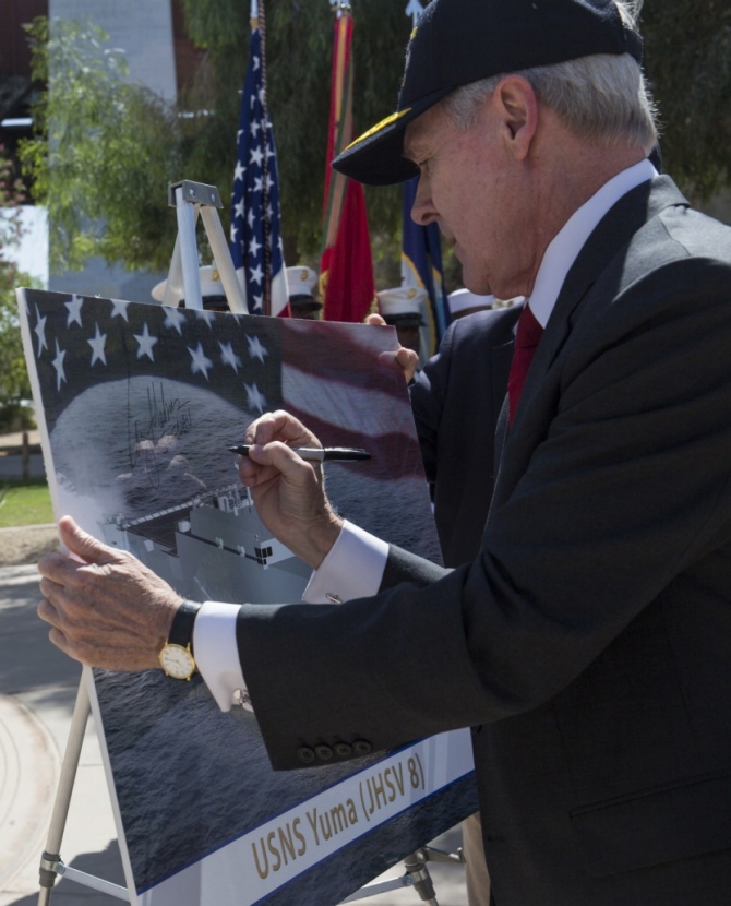 Secretary of the Navy Raymond E. Mabus Jr. signs a presentation commemorating the harmonious existence between the community of Yuma and its military installations, during the ship’s naming ceremony at Gateway Park in Yuma, Ariz., 25 April 2014. (Lance Cpl. James Marchetti, USMC, Marine Corps Air Station Yuma, Dept. of Defense Photograph 1000w_q95, Defense Video Imagery Distribution System).