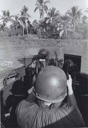 View from behind man wearing a helmet and firing a gun towards a river bank lined with palm trees