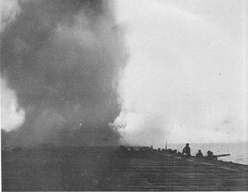 Photo 1: Looking aft on flight deck several seconds after hit showing column of water falling and smoke rising aft of flight deck. Prone figure in foreground is one of crew from starboard 40mm gun who was blown up and across deck. Firefighters and corpsmen are coming up from catwalk.
