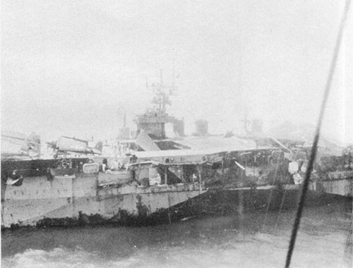 General view of forward damage from explosions in hangar. Note beam on flight deck crane, inclined forward elevator platform, hole blown in flight deck and panels blown out on the port side of the hangar.