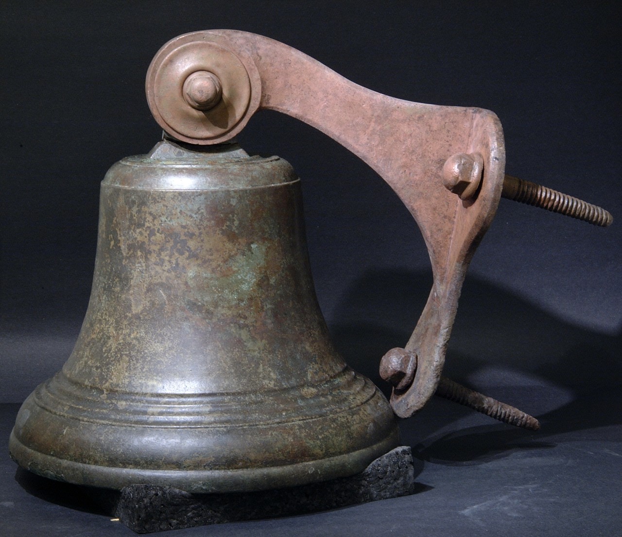 <p>A medium size bronze bell recovered from the CSS Alabama with a raised line at the top and three incised lines near the bottom rim of the bell. A bronze mounting racket is attached to the top of the bell which curves downward to a bronze plate with two screws. The mounting racket would have been screwed into the foremast allowing the bell to hang.</p>
