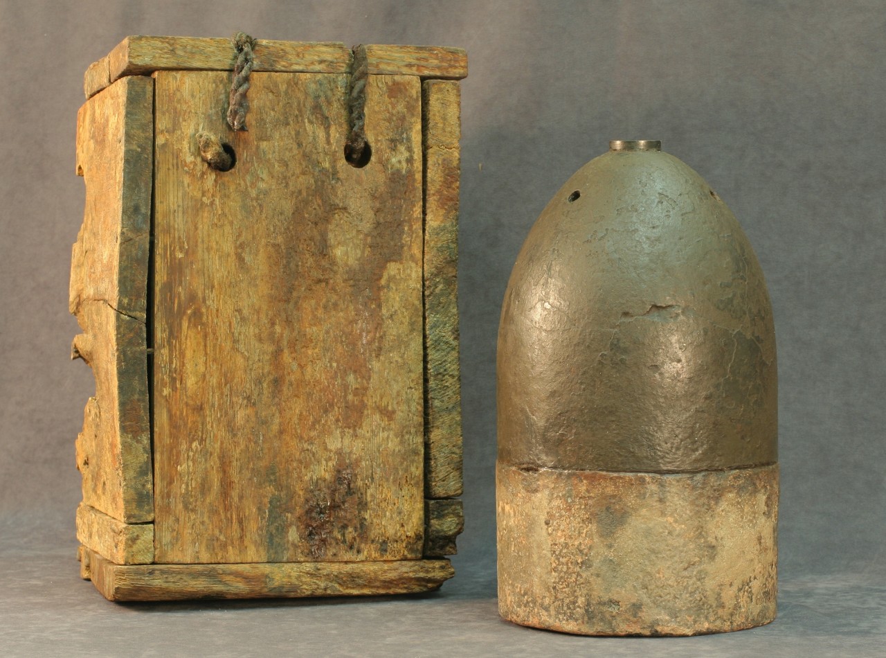 A round cast iron ammunition shell called a Bitten shell resting on a round wooden disc.  A brass fuse is located on the top of the shell. On either side of the shell are two square wooden containers that when put together make a box. The Britten shell was discovered inside this box resting on the wooden disc when it was recovered.  
