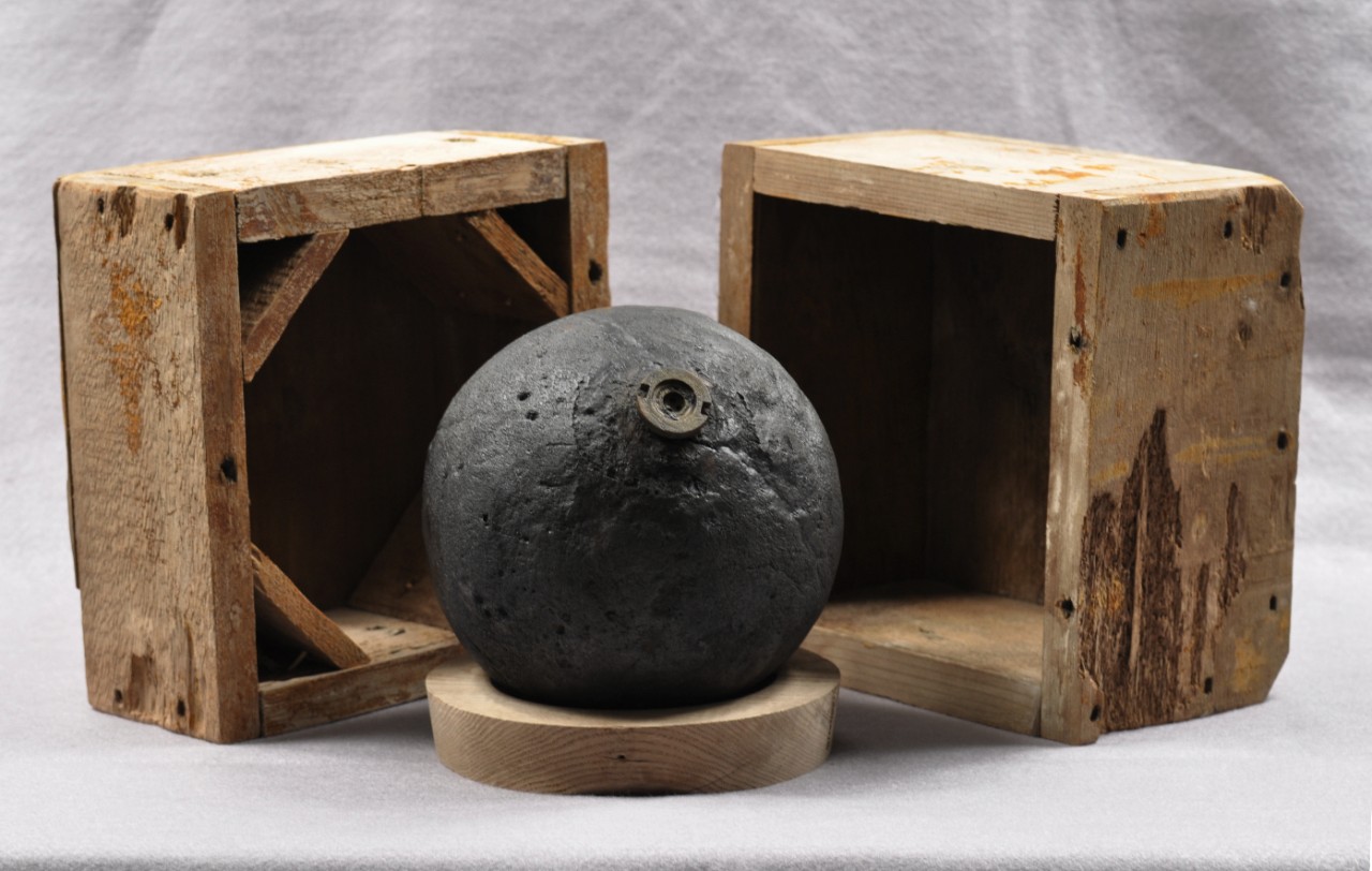 Cannon ball surrounded by a square wooden box.  The bottom section of the box is located on the left side of the cannon ball and the top section of the box is located on the right side of the cannon ball.  The cannon ball is resting on a circular wooden piece.