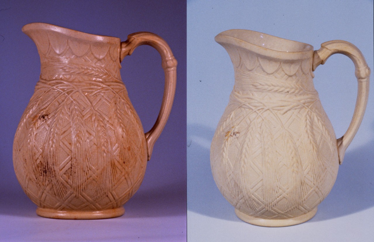 A tan colored ceramic pitcher recovered from CSS Alabama.  The rim or pouring lip of the pitcher is elongated with oval designs below the rim.  Twined wheat stems are around the narrow neck section with high relief wheat heads dangling downward on the body.  The bulbous body has a molded decoration with a double cross hatch design which tapers to a round base.  The handle is curved with a horizontal beaded band half way down the handle. The before and after conservation treatment are placed side by side. 