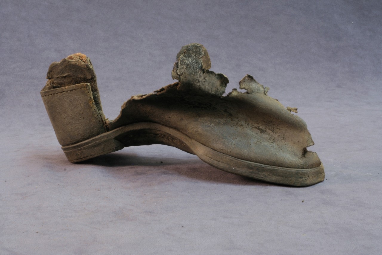 An incomplete left-footed leather shoe recovered from CSS Alabama.  The back section or heel cap and the front section is all that remains of the leather portion.  The leather is resting on the sole which is curved upward in the middle causing the shoe to be distorted.   